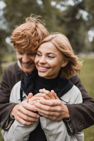 Heureux jeune homme en manteau échauffer les mains de petite amie gaie dans le parc automnal — Photo de stock