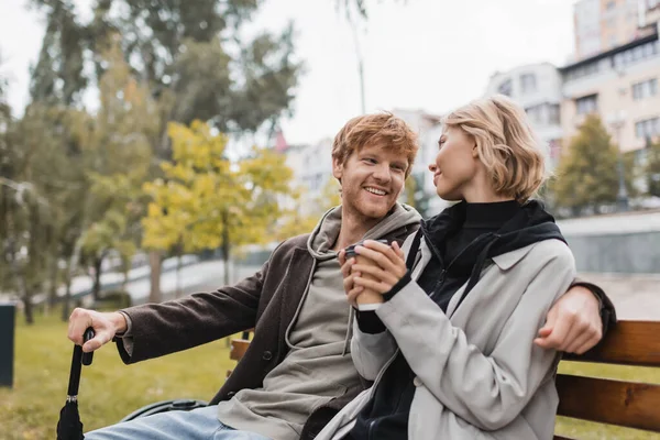 Heureux jeune homme en manteau regardant blonde copine tenant tasse en papier tout en étant assis sur le banc — Photo de stock