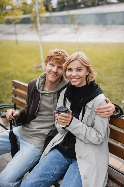 Jovem feliz no casaco abraçando namorada loira segurando copo de papel enquanto sentado no banco — Fotografia de Stock