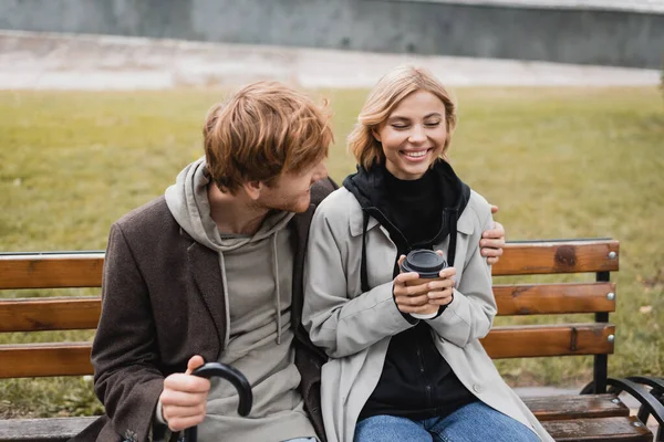 Joyeux jeune homme avec parapluie étreignant petite amie blonde tenant tasse en papier tout en étant assis sur le banc — Photo de stock