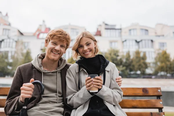 Happy young man with umbrella hugging girlfriend holding paper cup while sitting on bench — Stock Photo