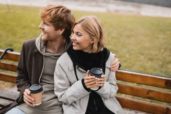 Feliz joven pareja abrazando y sosteniendo vasos de papel con café para ir mientras está sentado en el banco en el parque - foto de stock