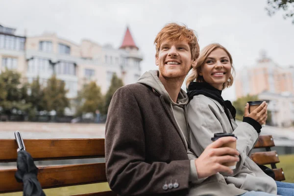 Fröhliches junges Paar umarmt und hält Pappbecher mit Coffee to go in der Hand, während es auf einer Bank im Park sitzt — Stockfoto
