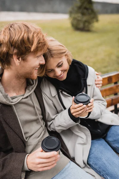 Sonriente pareja joven abrazando y sosteniendo tazas de papel con café para ir sentado en el banco en el parque — Stock Photo