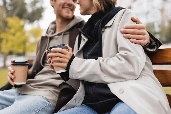 Vista recortada de feliz pareja joven abrazando y sosteniendo tazas de papel con café para ir en el parque - foto de stock