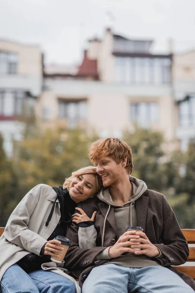 Щаслива молода жінка обіймає чоловіка і тримає каву, щоб піти, сидячи на лавці в парку — стокове фото
