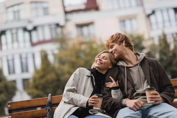 Sorridente giovane donna abbracciando l'uomo e tenendo il caffè per andare mentre seduto sulla panchina nel parco — Foto stock