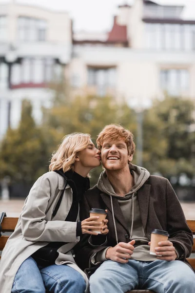 Sorrindo jovem mulher beijando bochecha do homem e segurando copo de papel enquanto sentado no banco no parque — Fotografia de Stock