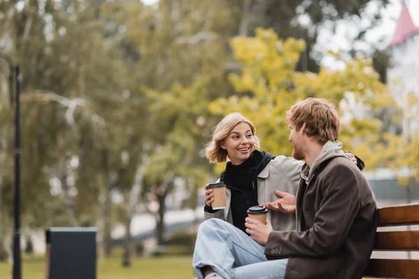 Sorrindo jovem casal conversando e segurando copos de papel com café para ir enquanto sentado no banco no parque — Fotografia de Stock