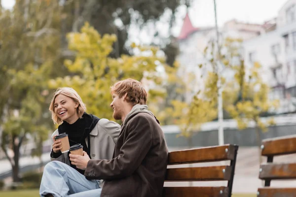 Alegre joven pareja riendo y sosteniendo vasos de papel con café para ir sentado en el banco en el parque - foto de stock