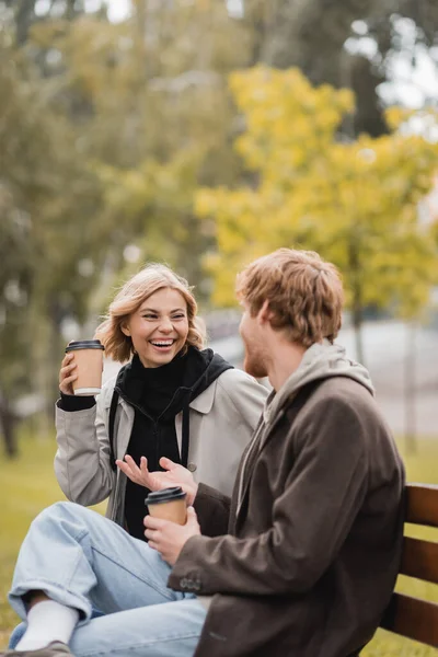 Feliz joven pareja riendo y sosteniendo vasos de papel con café para ir sentado en el banco en el parque - foto de stock
