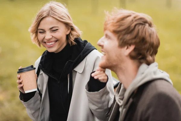 Mujer rubia feliz sosteniendo taza de papel y tocando la barbilla del novio pelirrojo - foto de stock