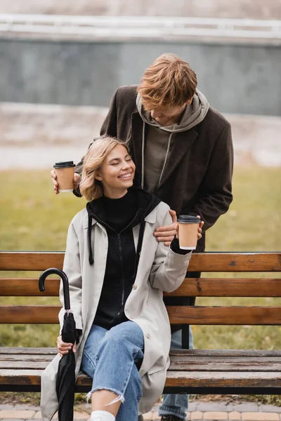 Uomo rosso che tiene tazze di carta vicino alla donna bionda seduta sulla panchina con ombrello nel parco — Foto stock