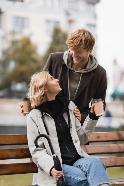 Rousse homme tenant des tasses en papier près de femme heureuse assis sur le banc avec parapluie dans le parc — Photo de stock