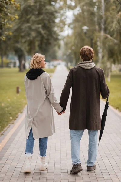 Back view of couple in coats holding hands and walking in autumnal park — Stock Photo