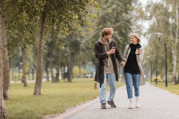 Full length of happy couple in coats holding hands and walking with coffee to go in autumnal park — Stock Photo