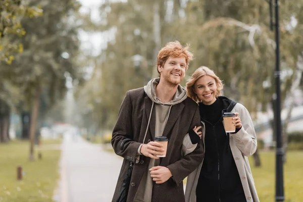 Feliz pareja joven en abrigos abrazando y caminando con copas de papel en el parque otoñal - foto de stock