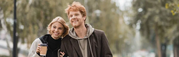 Happy young woman in coat hugging man and walking with paper cup in autumnal park, banner — Stock Photo