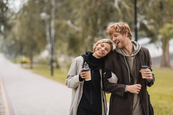 Alegre pareja joven en abrigos abrazando y caminando con vasos de papel en el parque otoñal - foto de stock