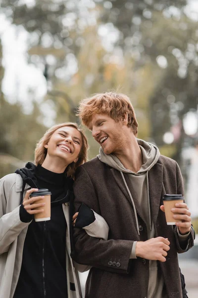 Позитивная молодая пара в пальто, обнимающая и гуляющая с бумажными стаканчиками в осеннем парке — стоковое фото