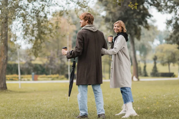 Longitud completa de joven feliz en abrigo sosteniendo taza de papel y de pie con la novia en el parque otoñal - foto de stock