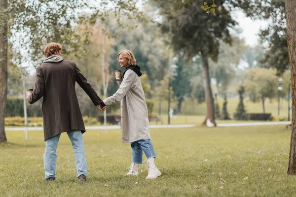 Voller Länge des rothaarigen jungen Mannes im Mantel mit Pappbecher und der Hand einer glücklichen Freundin im herbstlichen Park — Stockfoto