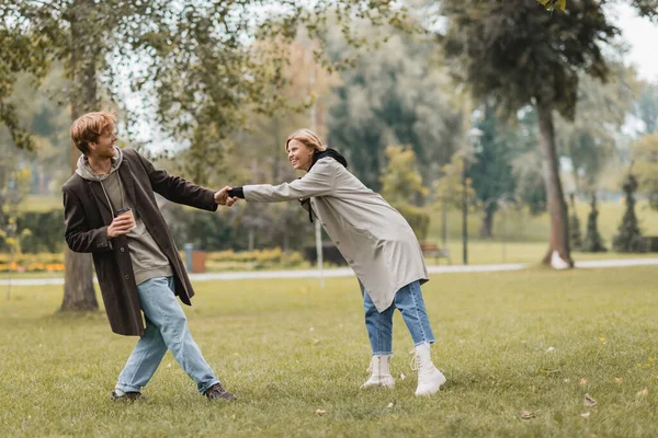 Полная длина счастливого молодого человека в пальто держа бумажную чашку и тянет подружку в осенний парк — стоковое фото