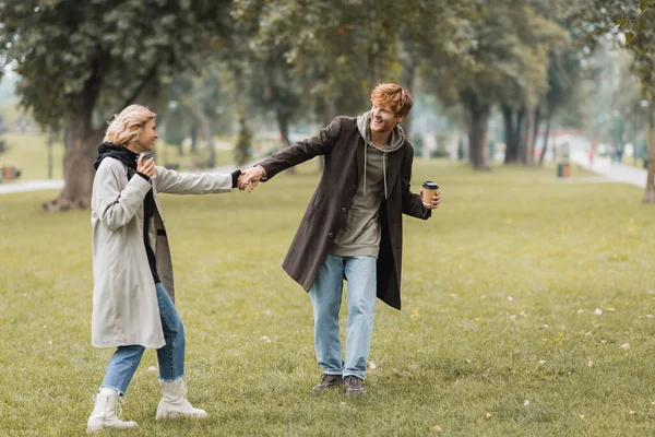 Full length of joyful man in coat holding hands with smiling girlfriend in autumnal park — Stock Photo