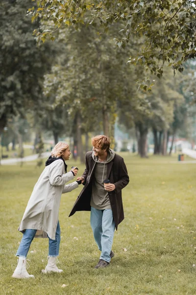 Full length of joyful man in coat holding paper cup and hand of pleased girlfriend in autumnal park — Stock Photo