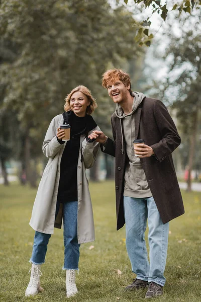 Full length of positive man with paper cup holding hands with pleased girlfriend in autumnal park — Stock Photo