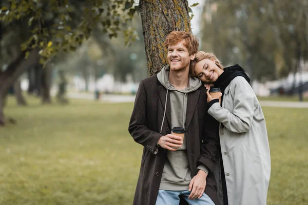 Happy blonde woman holding paper cup and leaning on shoulder of smiling boyfriend near tree trunk — Stock Photo