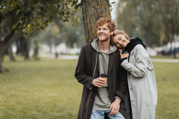 Glückliche blonde Frau hält Pappbecher in der Hand und stützt sich auf die Schulter ihres lächelnden Freundes in der Nähe des Baumstammes — Stockfoto