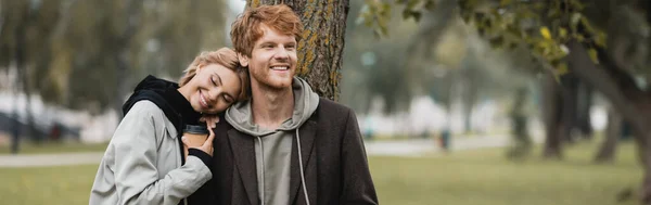 Mujer rubia feliz sosteniendo taza de papel y apoyándose en el hombro de novio sonriente cerca del tronco del árbol, pancarta - foto de stock