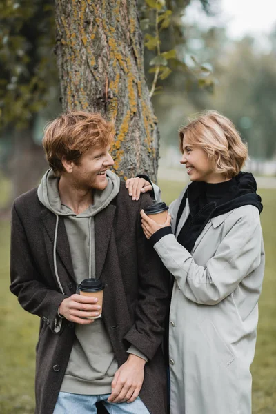 Glückliche blonde Frau hält Pappbecher in der Hand und lacht mit Freund in der Nähe von Baumstamm — Stockfoto