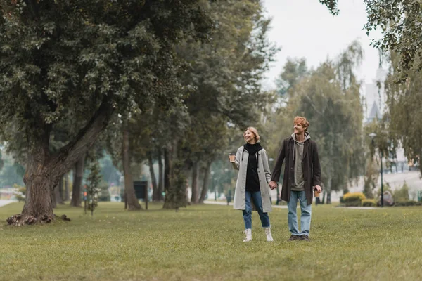 Full length of happy redhead man and cheerful woman holding coffee to go while walking in park — Stock Photo