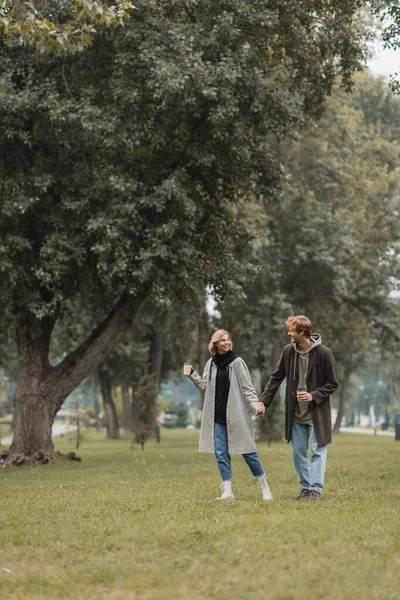 Pleine longueur de rousse positive homme et femme gaie tenant café pour aller tout en marchant dans le parc — Photo de stock