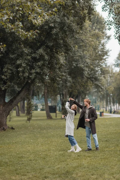 Полный рост счастливого рыжего мужчины и веселой женщины, держащей кофе, чтобы пойти на танцы в парке — стоковое фото