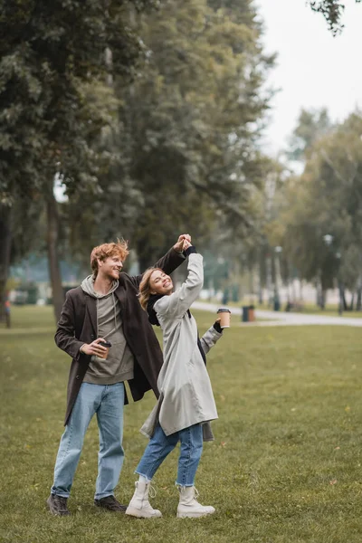 Полная длина счастливого рыжеволосого мужчины и веселой женщины, держащей кофе, чтобы пойти во время кручения во время танца в парке — стоковое фото