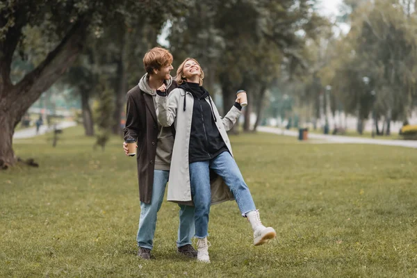 Comprimento total de homem ruiva feliz e mulher alegre segurando café para ir enquanto ri no parque — Fotografia de Stock