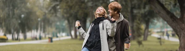 Happy redhead man looking at cheerful woman holding coffee to go while laughing in park, banner — Stock Photo