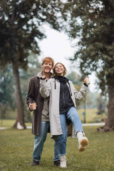 Volle Länge des glücklichen rothaarigen Mannes und der zufriedenen jungen Frau, die beim Spazierengehen im Park Kaffee zum Mitnehmen hält — Stockfoto