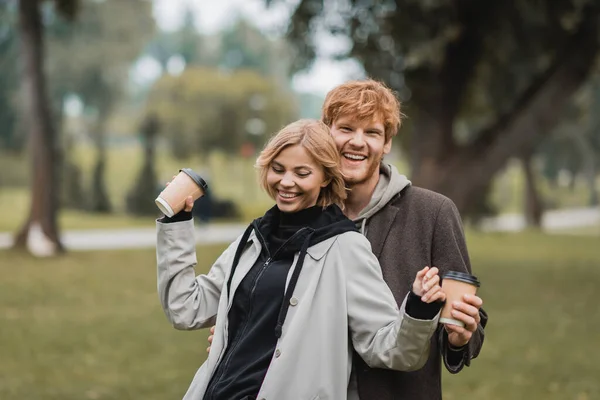 Heureux rousse homme et heureux jeune femme tenant café pour aller tout en marchant dans le parc — Photo de stock