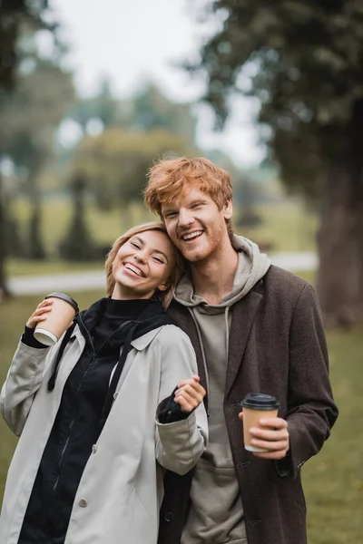 Alegre hombre y feliz joven mujer sosteniendo café para ir mientras se ríe en el parque - foto de stock