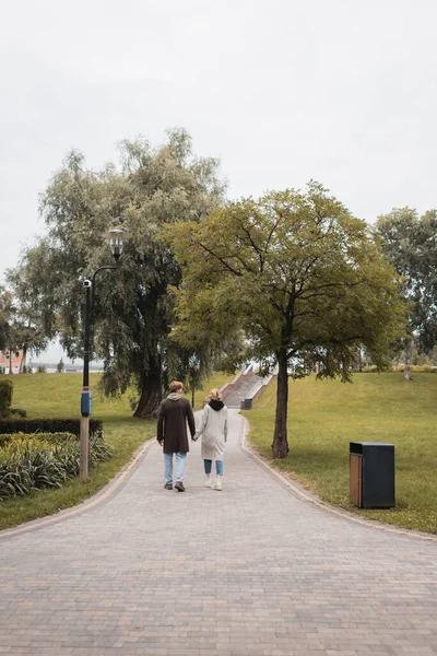 Vista trasera del hombre pelirrojo feliz y la mujer joven complacida cogida de la mano mientras camina en el parque - foto de stock