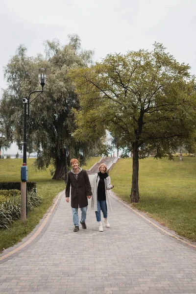 Full length of joyful redhead man and pleased young woman in coat holding hands while walking in park — Stock Photo