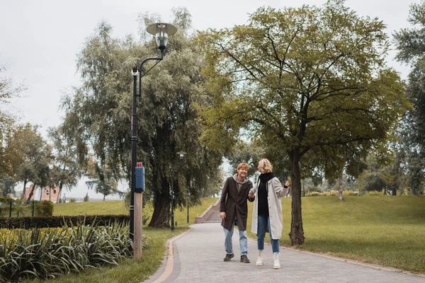 Volle Länge des glücklichen rothaarigen Mannes und der blonden jungen Frau im Mantel, die Händchen hält, während sie im Park spaziert — Stockfoto