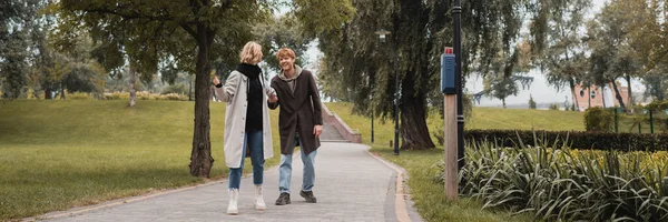 Повнометражний щасливий рудий чоловік і блондинка молода жінка в пальто тримає руки під час ходьби в парку, банер — стокове фото
