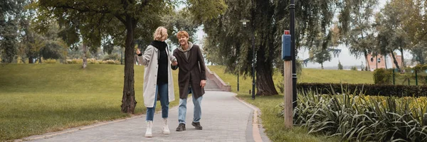 Comprimento total de homem ruivo feliz e mulher jovem loira de casaco de mãos dadas enquanto caminha no parque, banner — Fotografia de Stock