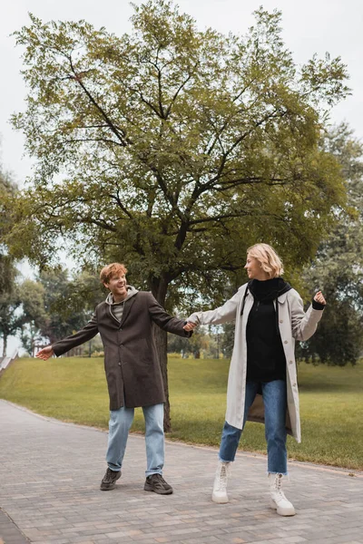 Longitud completa de hombre feliz y mujer complacida en abrigos tomados de la mano mientras camina en el parque - foto de stock