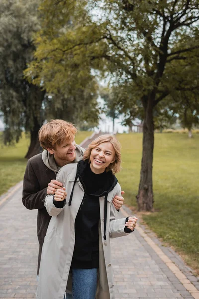Feliz hombre abrazando alegre joven mujer con los ojos cerrados riendo en el parque - foto de stock